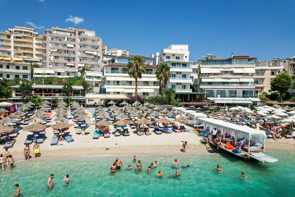Children and adults swimming in shallow turquoise water at a beach in Saranda, lined with straw umbrellas and seafront hotels